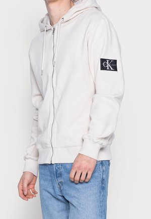 Mężczyzna w kremowej bluzie z kapturem zapinanej na suwak z naszywką z logo Calvin Klein na lewym rękawie i jasnoniebieskich dżinsach.
