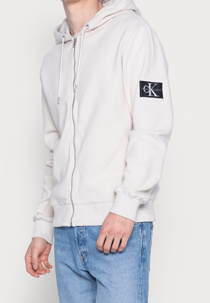 Homme portant un sweat à capuche zippé écru avec un patch logo Calvin Klein sur la manche gauche et un jean bleu clair.