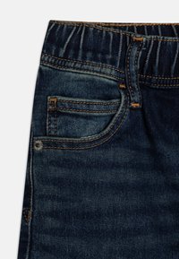 Närbild av mörkblå denimjeans med elastiskt midjeband, framficka och detaljer i kopparfärgad sömnad.