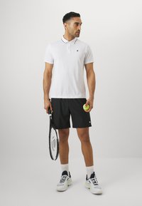 Weißes Poloshirt mit schwarzem Kragen, schwarze Shorts, hält einen Tennisschläger und einen Ball. Weiße Sportschuhe mit grauen Akzenten.