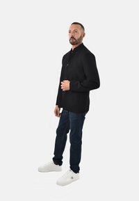 Giacca nera a maniche lunghe con colletto alto e chiusura a bottoni, abbinata a jeans blu scuro e sneaker bianche. Look semplice e sartoriale.