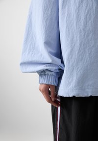 A person's hand rests at their side, wearing a light blue, oversized shirt with gathered sleeves, paired with black pants featuring a side stripe.