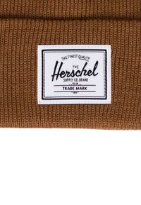 Berretto lavorato a maglia marrone con una patch bianca recante un'etichetta con testo nero, che mostra il nome del marchio "Herschel" e dettagli decorativi.