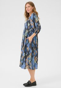 Vestido maxi de tela ligera que presenta un patrón abstracto multicolor con azules, marrones y verdes. Incluye bolsillos y mangas sueltas.
