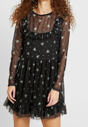 Femme portant une robe longue noire transparente à manches longues avec de petits motifs floraux blancs et des détails à volants sur la poitrine et l'ourlet.