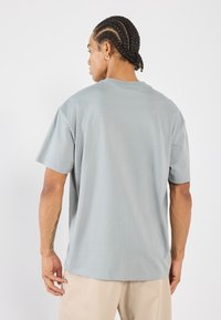 Světlé šedé tričko s krátkým rukávem, které má kulatý výstřih, volný střih a jemnou texturu. Na zadní straně je jednoduchý design bez potisků či log.
