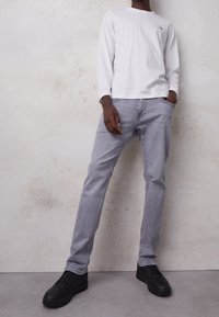 Camisa blanca de manga larga con logo, jeans grises y zapatillas negras. Los jeans tienen un corte recto; la camisa tiene un cuello redondo y puños acanalados.