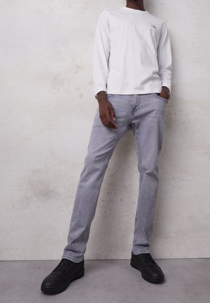 Camisa blanca de manga larga con logo, jeans grises y zapatillas negras. Los jeans tienen un corte recto; la camisa tiene un cuello redondo y puños acanalados.