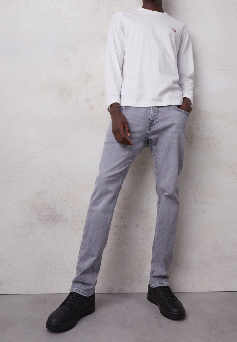 Camisa blanca de manga larga con logo, jeans grises y zapatillas negras. Los jeans tienen un corte recto; la camisa tiene un cuello redondo y puños acanalados.
