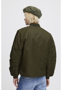 Veste bomber vert olive avec col côtelé, manches froncées et poches latérales, portée avec un béret en laine assorti pour une touche supplémentaire.
