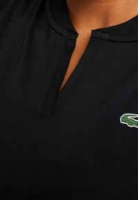 Svart skjortahalssnitt med en liten V-formad öppning och en grön krokodil-logotyp broderad på höger sida.