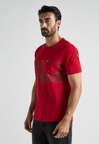 Rode katoenen t-shirt met een borstzak en een textuur grafisch ontwerp in een lichtere tint over de voorkant. Korte mouwen en een ronde hals.