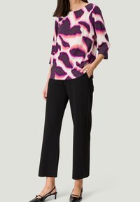 Bunt gemusterte Bluse in Lila, Pink und Weiß, kombiniert mit schwarzen, weit geschnittenen Hosen. Sie hat 3/4-Ärmel und einen runden Halsausschnitt.