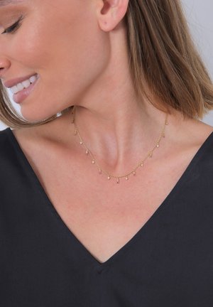 Goldene Kettenhalskette mit tropfenförmigen rosa und weißen Edelsteinen, die zarte hängende Akzente aufweist. Getragen von einem Modell mit einem V-Ausschnitt-Oberteil.