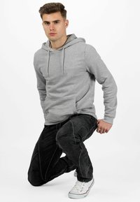 Grijze hoodie met een voorzak, koordjes en geribbelde manchetten, gecombineerd met zwarte jeans met contrasterende stiksels en witte sneakers.