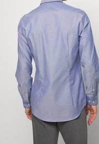 Camicia button-up blu chiaro con maniche lunghe, colletto standard e un motivo testurizzato sottile. Caratterizzata da un orlo curvo e polsini singoli.