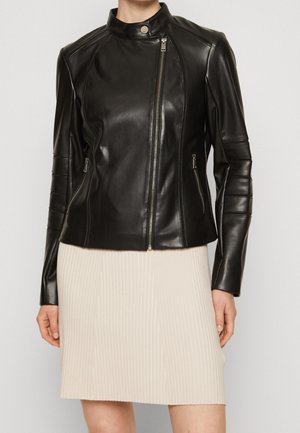 Veste en cuir noire ajustée avec une fermeture éclair asymétrique sur le devant et un col montant, portée avec une jupe plissée beige clair.