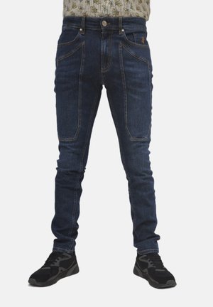 Jeans in denim blu scuro con vestibilità slim, tasche frontali e dettagli con doppio cucitura. Indossati con scarpe sportive nere.