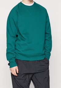 Modrozelená mikina s kulatým výstřihem, žebrovanými manžetami a lemem. Má hladkou texturu a volný střih, doplněná tmavými kalhotami.