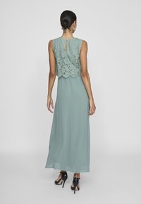 Robe longue vert doux avec un corsage en dentelle et un dos avec un détail en forme de trou. Tissu lisse et jupe longueur cheville. Chaussures à talons noires.