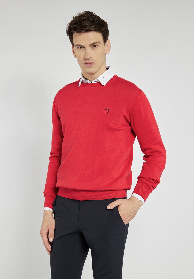 Czerwony sweter z miękkiego dzianinowego materiału, z okrągłym dekoltem, prążkowanymi mankietami oraz małym logo po lewej stronie. Nosi się go na koszuli z kołnierzykiem.