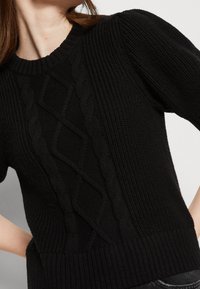 Pull en tricot noir avec un motif de câble texturé, col rond à côtes et manches courtes bouffantes. Tissu doux et épais, coupe décontractée.