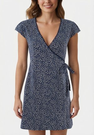 Mujer con vestido cruzado azul marino con pequeño estampado floral blanco y un lazo lateral, de pie con los brazos relajados contra un fondo claro liso.