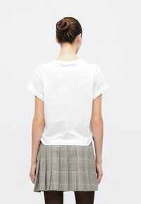 Frau mit Hochsteckfrisur, die ein weißes Kurzarmshirt und einen plissierten grauen Karorock trägt, vor einem schlichten weißen Hintergrund stehend.