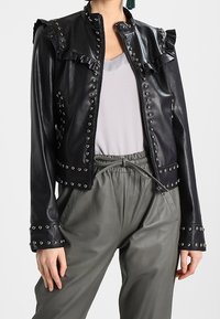 Veste en cuir noire avec col froncé, clous argentés le long des coutures et coupe ajustée, associée à un pantalon gris décontracté.