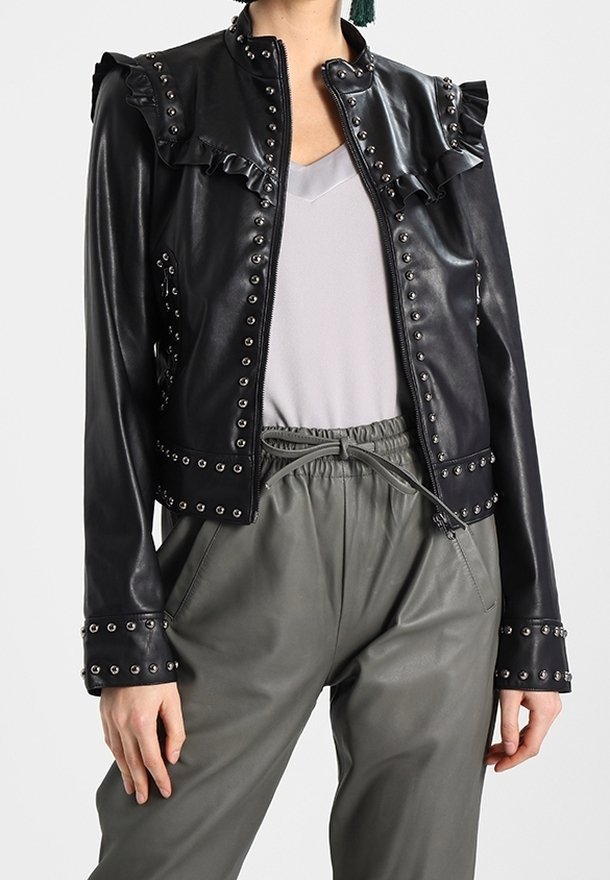 Veste en cuir noire avec col froncé, clous argentés le long des coutures et coupe ajustée, associée à un pantalon gris décontracté.