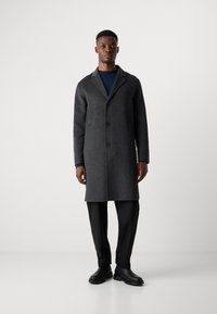 Grijze wollen jas met ingeklapte kraag, drie knopen en zijzakken, gedragen over een marineblauwe trui en zwarte broek, gecombineerd met zwarte schoenen.