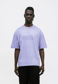 Μπλουζάκι από βαμβάκι σε χρώμα λεβάντας με υπερμεγέθη εφαρμογή, εμφανίζοντας το τολμηρό, ανάγλυφο λογότυπο "HUGO" στο μπροστινό μέρος. Κοντά μανίκια και στρογγυλή λαιμόκοψη.