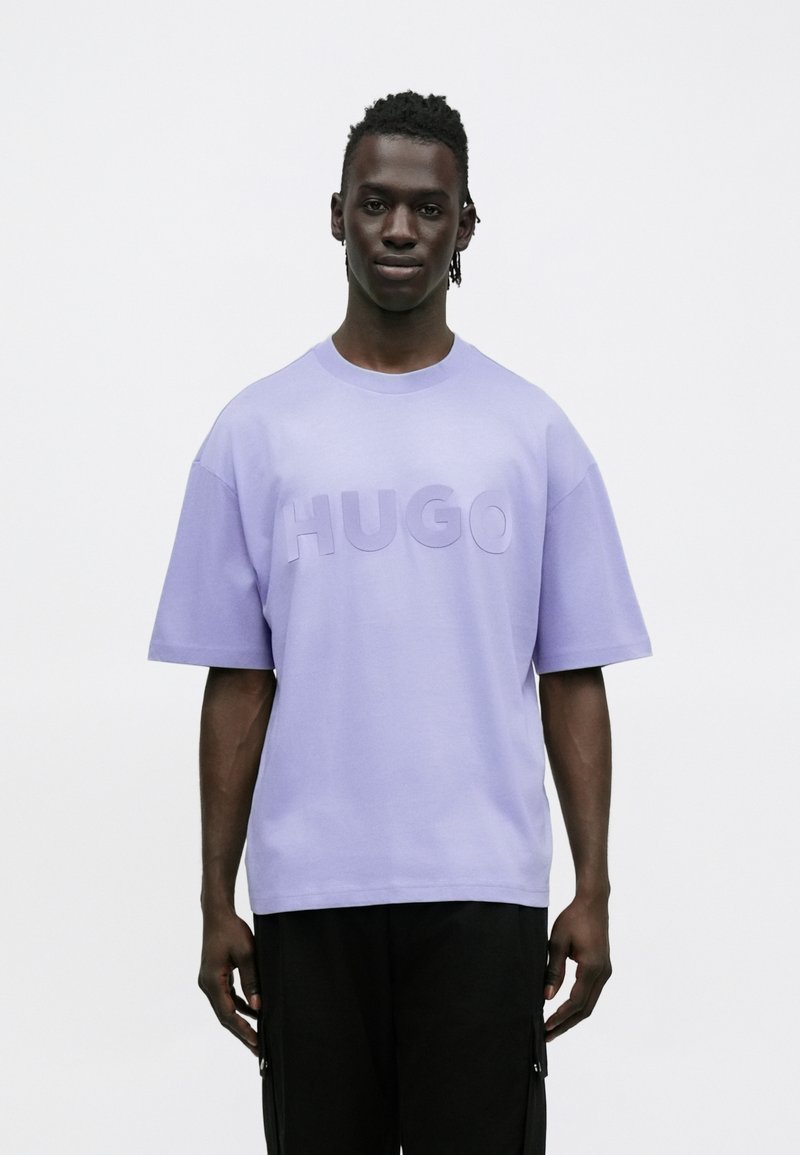 Μπλουζάκι από βαμβάκι σε χρώμα λεβάντας με υπερμεγέθη εφαρμογή, εμφανίζοντας το τολμηρό, ανάγλυφο λογότυπο "HUGO" στο μπροστινό μέρος. Κοντά μανίκια και στρογγυλή λαιμόκοψη.