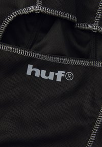 Zwart sportstof met een meshstructuur, voorzien van grijze "huf" branding en contrasterende witte stiksels langs de naden.