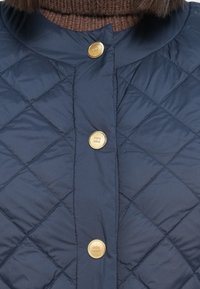 Marineblå quiltet jakke med rund krage, dekorert med gullknapper og en strukturert brun strikkekrage under.