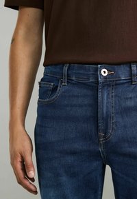 Jeans de mezclilla azul oscuro con un diseño ajustado, que incluyen un bolsillo frontal, costuras de cobre y un cierre de botón de metal en la cintura.