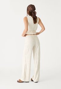 Blouse beige clair sans manches avec un lien à nouer dans le dos, associée à un pantalon à jambes larges orné d'un motif floral en relief. Foulard marron à pois.