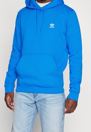 Mężczyzna noszący jasnoniebieską bluzę z kapturem Adidas z przednią kieszenią i jasnoniebieskie dżinsy, stojący na jednolitym tle.