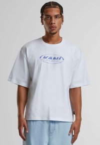 White oversized T-shirt made of cotton with a round neckline. Features a blue logo print, short sleeves, and a relaxed fit.