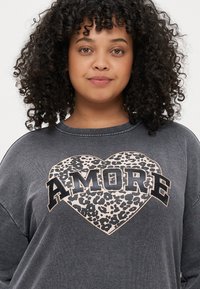 Sweatshirt cinza com um decote arredondado; apresenta uma grafismo de coração com padrão de leopardo com "AMORE" em letras pretas em destaque. Cabelo encaracolado emoldurando o rosto.