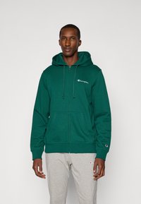 Groene rits-hoodie van zachte stof met een voorzak, verstelbare trekkoordcapuchon en geborduurd logo op de borst. Grijze joggingsbroek.