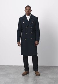 Marineblå dobbeltradet uldfrakke med gyldne knapper, brede revers og længere pasform, kombineret med mørke bukser og brune støvler. Tekstureret stof.