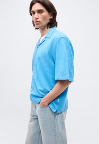 Νέος άνδρας με μεσαίου μήκους σκούρα μαλλιά φοράει ανοιχτό γαλάζιο πουκάμισο με κοντά μανίκια και τζιν σε ανοιχτή απόχρωση, στέκεται με το ένα χέρι στην τσέπη.