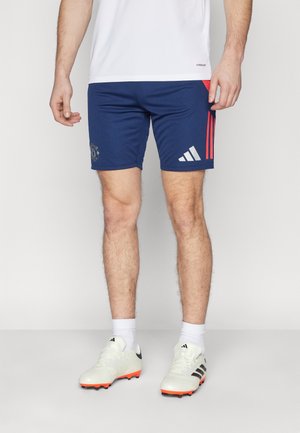 Homme portant un short de sport Adidas bleu avec des bandes rouges, des chaussettes blanches et des crampons Adidas blancs avec des bandes noires.