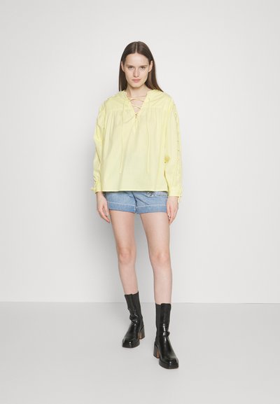 Haut à manches longues jaune avec un décolleté lacé, associé à un short en jean. Le mannequin porte des bottines noires à talons. Fond minimaliste.