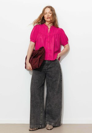 Femme aux longs cheveux portant une blouse rose vif, un jean à jambes larges gris foncé, des chaussures à imprimé léopard, et tenant un sac à motifs contre un mur blanc.