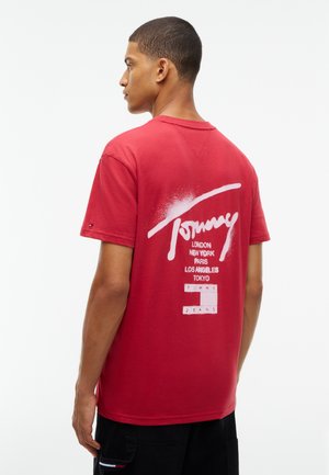 Млад мъж, гледащ настрани, носещ червена тениска Tommy Jeans с имена на градове, отпечатани на гърба с бял текст.