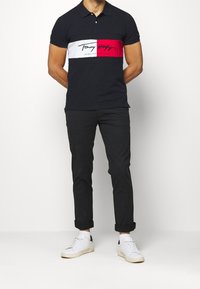 Polo shirt de un oscuro azul marino con una franja horizontal blanca y roja que presenta el logo "Tommy Hilfiger". Combinado con pantalones negros y zapatillas blancas.