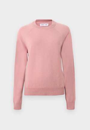 Myk rosa genser med rund hals og lange ermer, laget av strikket materiale med ribbestrikket mansjetter og hem, vist mot en lys, ensfarget bakgrunn.