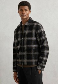 Sort og gråternet uldjakke med lynlås foran, to frontlommer og krave. Båret over en brun sweater og sorte bukser.